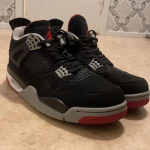 Jordan 4 Bred - Snygga svarta Jordan 4 sneakers med grå och röda detaljer. Skorna har snörning och en klassisk Nike Air-logga på hälen. Perfekta för en sportig look. Använd endast två gånger, kommer inte med originalboxen tyvärr.