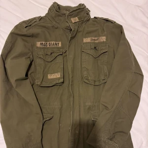 Brandit militärinspirerad jacka - Säljer en olivgrön militärinspirerad jacka med flera fickor och detaljerade broderier. Jackan har en hög krage och långa ärmar, perfekt med Skjorta eller T-shirt under.