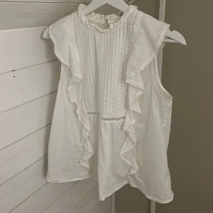 Vit ärmlös blus med volangdetaljer - Säljer en söt vit blus med volangdetaljer framtill och baktill. Blusen är ärmlös och har knäppning i nacken. Använd fåtal gånger, köpt i våras. Pris kan diskuteras