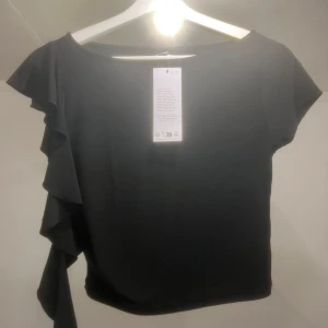 Svart topp med volangdetalj från GinaTricot - Snygg svart topp från GinaTricot med en unik volangdetalj på ena sidan. Toppen har korta ärmar och en rund halsringning. Perfekt för en stilren look. Aldrig använd