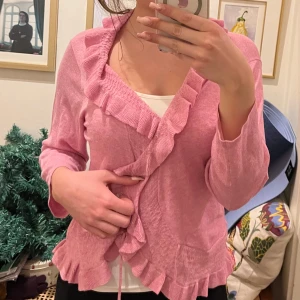 Rosa volangkofta - Säljer en söt rosa kofta med volangdetaljer. Koftan har långa ärmar och en fin omlott-design som ger en feminin touch. Perfekt för att lägga till lite färg i garderoben!