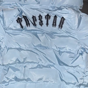 Ljusblå pufferjacka från Trapstar - Snygg ljusblå pufferjacka från Trapstar med deras ikoniska logga på ryggen. Jackan har en dragkedja framtill och en hög krage för extra värme. Perfekt för kyliga dagar.