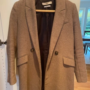 Beige/ljusbrun kappa från Mango - Ljusbrun kappa från Mango med klassisk design och knappar framtill. Perfekt för höst och vår, med en stilren look som passar till många tillfällen. I slitet/använt skick.