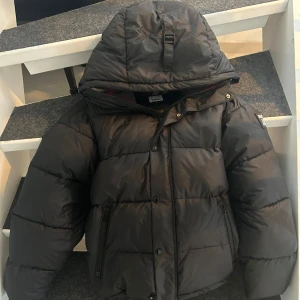 Svart dunjacka från Burberry - Säljer en snygg svart dunjacka från Burberry med huva och dragkedja. Jackan har en broderad logotyp på bröstet och är perfekt för kalla dagar. Den har en quiltad design och är både stilren och funktionell. kan även bli till väst!