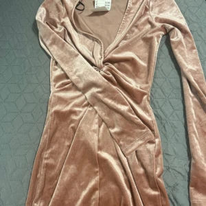 Rosa sammet klänning från H&M - Snygg rosa sammet klänning från H&M med långärmad design och v-ringad hals. Klänningen har en knutdetalj framtill som ger en elegant touch. Perfekt för en stilfull look.