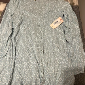 Blå blommig klänning från Nelly - Säljer en söt blå blommig klänning från Nelly med långa ärmar och v-ringning. Perfekt för en romantisk look med sitt fina mönster och bekväma passform. Klänningen har knappar framtill och är i storlek EU 36.