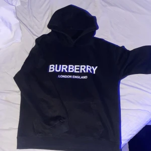 Svart Burberry Hoodie - Snygg svart hoodie från Burberry med vit text 'BURBERRY LONDON ENGLAND' på framsidan. Perfekt för en stilren och bekväm look. Den har en klassisk huva och ribbade muddar. Helt nytt skick, storlek M. Hör av er!😁