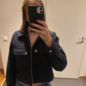 Svart bomberjacka med dragkedja - Snygg svart bomberjacka med dragkedja och knappar framtill. Jackan har en croppad passform och långa ärmar. Perfekt för en trendig look.