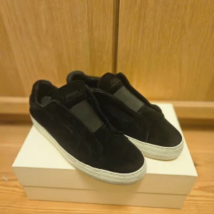 Svarta sneakers från Axel Arigato - Snygga svarta sneakers från Axel Arigato i mocka med vit sula. Perfekta för en stilren look. De har en minimalistisk design utan snörning, vilket gör dem lätta att ta på och av. Fraktar inom 24h