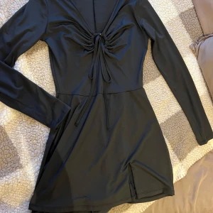 Svart playsuit, S - Snygg svart playsuit som ska se ut som klänning. Med långa ärmar och en ring i mitten av bröstet som man kan knyta. Klänningen har en slits i ena sidan. Knappt använd. Storlek S. från new Yorker.