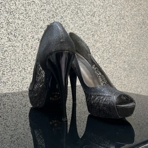 Svarta pumps med spetsdetaljer - Eleganta svarta pumps med hög klack och spetsdetaljer. Skorna har en öppen tå och en glansig finish som ger en lyxig känsla.