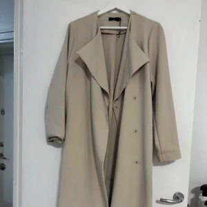 Beige trenchcoat med bälte - Snygg beige trenchcoat med knappar och bälte i midjan. Perfekt för höst och vår. Långärmad design med klassisk krage. 