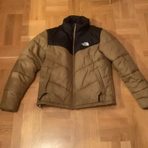 Brun och svart pufferjacka från The North Face - Säljer en snygg brun och svart pufferjacka från The North Face. Jackan har en dragkedja framtill och är långärmad. Perfekt för kyligare dagar med sin quiltade design. Skick 9/10, använd ett fåtal gånger. 
