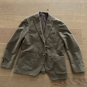 Beige kavaj från Massimo Dutti - Snygg beige kavaj från Massimo Dutti i bomull med en touch av elastan för extra komfort. Kavajen har klassiska slag, två knappar och praktiska fickor framtill. Perfekt för en stilren look.