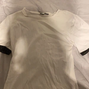 Vit t-shirt med glittriga detaljer från Zara - Säljer en stilren vit t-shirt från Zara med korta ärmar och glittriga detaljer vid ärmsluten. 