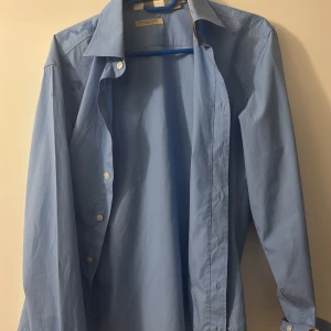 Blå skjorta från Burberry Brit - Snygg blå skjorta  Burberry Brit med klassisk krage och knappar framtill. Skjortan har långa ärmar och en diskret broderad logga på bröstet. Perfekt för en stilren look. Pris kan diskuteras 