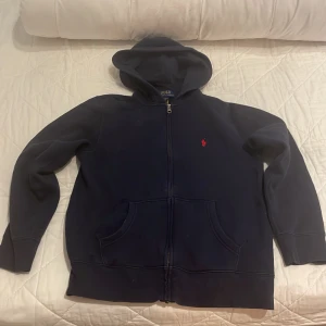  Polo Ralph Lauren zip  - Säljer en snygg mörkblå hoodie från Polo Ralph Lauren med dragkedja och huva. Den har en liten röd logga på bröstet och praktiska fickor framtill. Perfekt för en avslappnad stil. Barnstorlek: L Vuxen: Xs 