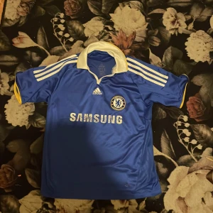 Blå Chelsea fotbollströja från Adidas - Säljer en blå Chelsea fotbollströja från Adidas med korta ärmar och vita detaljer. Tröjan har klubbens emblem och Samsung-logga på framsidan. Perfekt för fans av Chelsea FC! 💙