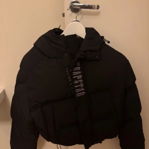 Svart pufferjacka från Trapstar - Snygg svart pufferjacka från Trapstar med huva och tryck både fram- och baksidan. Använd ett fåtal gånger. Ny pris 4400kr. Storlek Xs