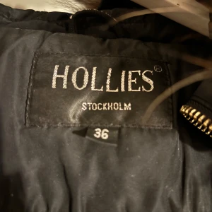Svart dunjacka från Hollies - Snygg svart dunjacka från Hollies med pälskrage och dragkedja. Jackan har flera praktiska fickor med dragkedjor och en quiltad design som ger en stilren look. Perfekt för kyliga dagar.