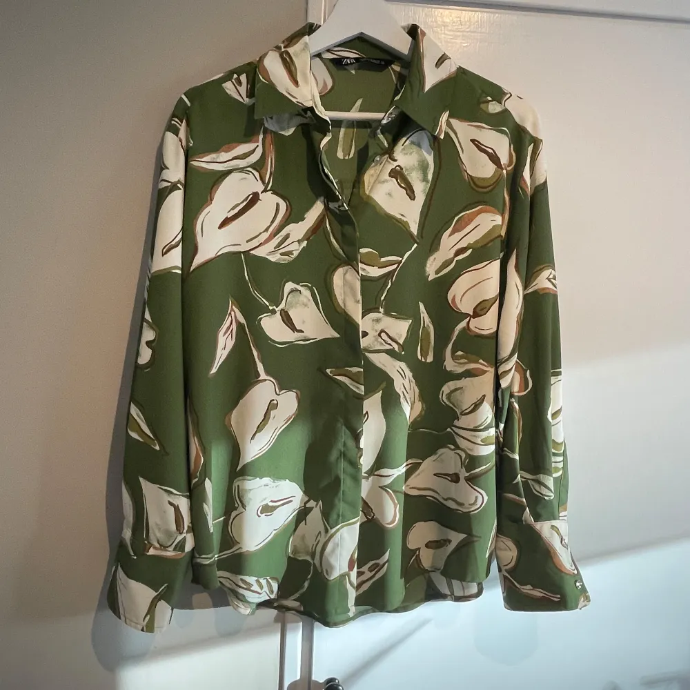 Snygg grön skjorta från Zara med ett stort blommönster i vitt och brunt. Skjortan har långa ärmar och en klassisk krage. Perfekt för en stilren look.. Paidat.