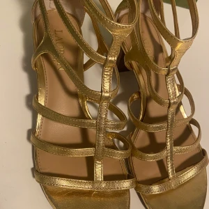 Guldiga sandaler från Ralph Lauren - Eleganta guldiga sandaler från Ralph Lauren med dragkedja bak och flera remmar. Perfekta för en glamorös look.