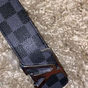 Rutig bälte från Louis Vuitton - Snyggt bälte från Louis Vuitton med ett klassiskt rutigt mönster i svart och grått. Bältet har en stilren silverfärgad spänne med märkets logotyp. Perfekt för att ge en lyxig touch till din outfit. Pris kan diskuteras genom snabb affär 