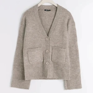 Beige kofta med knappar - Mysig beige kofta med knappar framtill och två praktiska fickor. Den har en ribbad nederkant och muddar, vilket ger en stilren look. Perfekt för kyliga dagar.