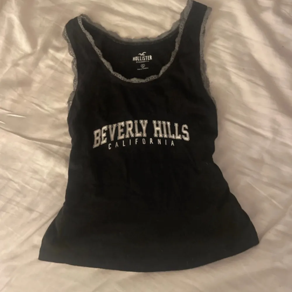 Snygg svart linnetopp från Hollister med texten 'Beverly Hills California' i vitt på framsidan. Toppen har spetsdetaljer runt halsringningen och ärmlösa kanter. Perfekt för en avslappnad stil.. Yläosat.