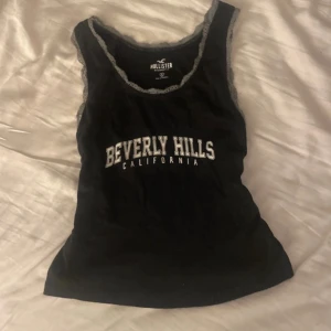 Svart linnetopp från Hollister - Snygg svart linnetopp från Hollister med texten 'Beverly Hills California' i vitt på framsidan. Toppen har spetsdetaljer runt halsringningen och ärmlösa kanter. Perfekt för en avslappnad stil.