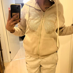 Beige huvtröja med fjärilsdetalj - Säljer en beige huvtröja med glittrande fjärilsdetalj på framsidan. Tröjan har dragkedja och långa ärmar, perfekt för en avslappnad stil. Den har en bekväm passform och är idealisk för vardagsbruk.