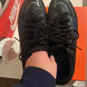 Svarta fotbollsskor från Nike - Snygga svarta fotbollsskor från Nike med blå detaljer och snörning. Skorna har en slitstark sula med dobbar, perfekt för spel på gräsplaner. De är designade för att ge bra grepp och stabilitet under matchen.