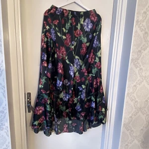 Blommig kjol från Visual Clothing Project - Snygg långkjol med blommigt mönster i rött, lila och grönt på en svart bakgrund. Kjolen har volangdetaljer och en elastisk midja för extra komfort. Perfekt för att ge en färgglad touch till din outfit. Asymmetrisk så längre baktill 