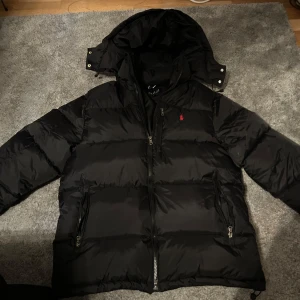 Svart dunjacka från Ralph Lauren - Säljer en svart dunjacka från Ralph Lauren med röd broderad logga på bröstet. Jackan har en praktisk huva och dragkedja framtill samt två fickor med dragkedjor. Perfekt för kyliga dagar!