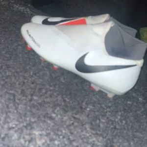 Nike phantom fotbollsskor  - Snygga vita fotbollsskor från Nike med svarta detaljer och en röd insida. Skorna har en strömlinjeformad design och är perfekta för fotbollsplanen. Diskuterar pris
