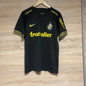 Svart AIK fotbollströja med gula detaljer L - Snygg svart AIK fotbollströja från Nike med gula detaljer och logotyper. Tröjan har korta ärmar med ett mönster på ärmsluten. Perfekt för alla AIK-fans! 