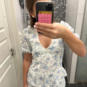Blommig klänning med volangdetaljer - Charmig vit klänning med blå blommönster och volangdetaljer. Klänningen har korta ärmar och en v-ringad design. Perfekt för somriga dagar med sin lätta och luftiga stil😍