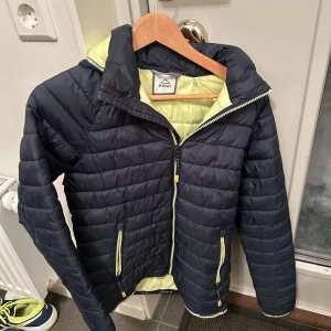 Svart och grön pufferjacka från McKinley - Säljer en svart pufferjacka från McKinley med en fräsch grön insida. Jackan har dragkedja och två praktiska fickor framtill. Perfekt för kyliga dagar och ger en sportig look.