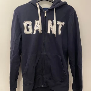 Mörkblå hoodie från GANT - Säljer en snygg mörkblå hoodie från GANT med dragkedja och stor logga på bröstet. Tröjan har en justerbar huva med vita snören och praktiska fickor framtill. Perfekt för en avslappnad stil. Skriv om ni har funderingar och priset är inte hugget i sten!