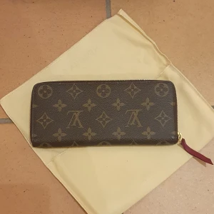 Plånbok från Louis Vuitton (PF.CLEMEMCE MONOG. FUCH.) - ❗️Skriv innan köp❗️ Säljer en elegant plånbok från Louis Vuitton i model PF.CLEMEMCE MONOG. FUCH. Har klassiskt monogrammönster. Plånboken har en dragkedja och flera fack inuti för kort och kontanter. Den är brun med en röd insida. Har används men omhändertagen. Kvitto och låda inkluderat.