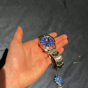 Blå klocka med röd och blå bezel - Snygg klocka med blå urtavla och en iögonfallande röd och blå bezel. Armbandet är i silverfärgad metall och klockan har en datumvisning. Perfekt för den som gillar en sportig och färgglad stil.