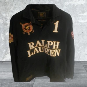 Svart pikétröja från Ralph Lauren - Snygg svart pikétröja från Ralph Lauren med broderade detaljer och nummer 1 på både fram- och baksida. Tröjan har långa ärmar och en klassisk krage. Perfekt för en stilren look.