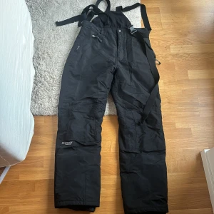 Svarta skidbyxor från Everest - Svarta skidbyxor från Everest med hängslen och dragkedja. Perfekta för vinterns kalla dagar. Byxorna har praktiska fickor med dragkedja och justerbara axelband för bästa passform. Endast använda 2 gånger . Nypris när jag köpte de var 1299:-.