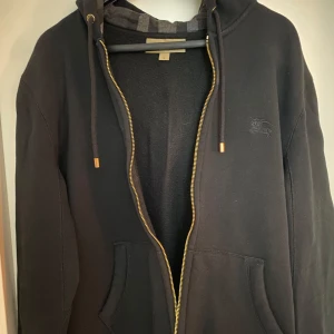 Burberry ZIP UP HOODIE (KOFTA) - Säljer en stilren svart zip up hoodie från Burberry. Klassiskt monogram i Luvan. Storlek M. Skick: 7/10 (Sparsamt använd). Kan mötas upp i Göteborg eller i närheten av Göteborg. Pris kan diskuteras vid snabb affär.