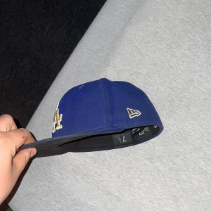 Blå keps från New Era - Snygg blå keps från New Era med LA-logga i guld framtill. Kepsen har en platt skärm och är en del av 59FIFTY-serien. Perfekt för att ge din outfit en sportig touch.
