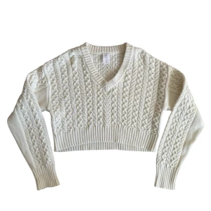 Vit stickad croppad tröja - Säljer en snygg vit stickad tröja med v-ringning och ett fint flätmönster. Tröjan är croppad och har långa ärmar med ribbade muddar. Endast använd vid ett tillfälle och är i bra skick!