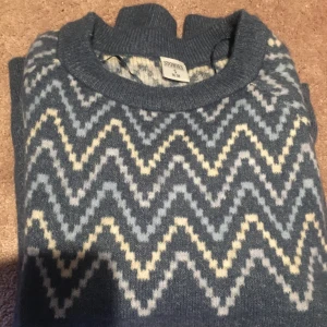 Blå stickad tröja från Stories - Säljer en snygg blå stickad tröja från Stories med ett zickzackmönster i vitt och beige. Tröjan har långa ärmar och en rund halsringning. Perfekt för kyliga dagar!