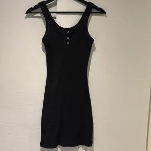 Svart ribbad klänning från Ambar - Snygg svart ribbad klänning från Ambar i medium. Den är ärmlös med en tight passform och har knappar framtill. Perfekt för en stilren look.