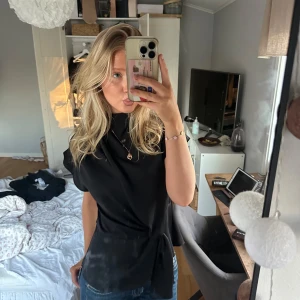 Svart knytblus - Säljer en elegant svart blus med knytning i midjan. Blusen har korta ärmar och en hög krage, vilket ger en stilren look. Perfekt för att klä upp en vardagsoutfit eller för en kväll ute.