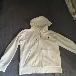 Vit hoodie med dragkedja från Ralph Lauren - Säljer en stilren vit hoodie från Ralph Lauren med dragkedja och broderad logotyp på bröstet. Tröjan har en bekväm passform med långa ärmar och en praktisk huva. Skick 7/10 lite nopprig med det går att få bort! Bara att höra av sig vid frågor!